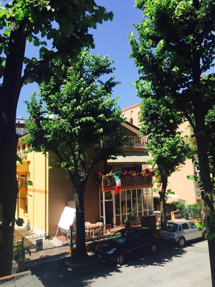 A serene European street scene framed by lush green trees showcases a charming yellow building with balconies adorned with flowers and an Italian flag, ideal for travelers seeking quaint urban retreats.
