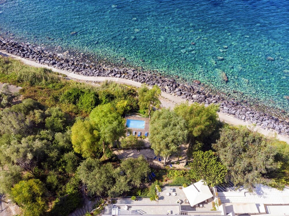 An aerial view captures a serene coastal retreat with lush greenery, a tranquil pool, and crystal-clear turquoise waters, ideal for a relaxing beachside vacation.