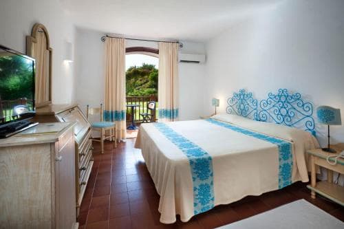 A cozy hotel room with a neatly made bed adorned in blue patterns, a balcony view of lush greenery, and warm terracotta flooring invites travelers seeking relaxation and comfort.
