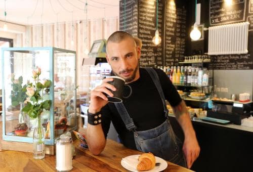 A cozy café scene features a man enjoying coffee and a croissant at a wooden counter, surrounded by charming decor like chalkboard menus and glass display cases, ideal for travelers seeking a relaxing breakfast spot.