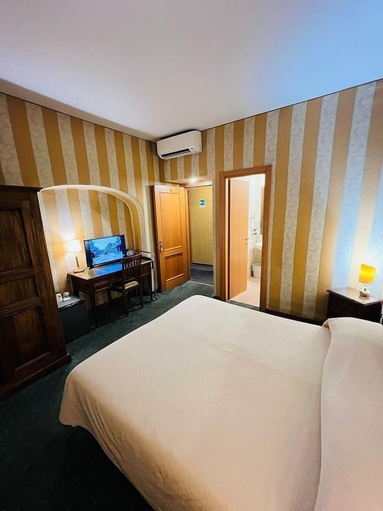 A cozy hotel room with striped wallpaper, a neatly made bed, a desk with a TV, and warm lighting creates an inviting atmosphere ideal for travelers seeking comfort and relaxation.