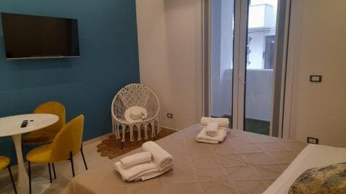 A cozy hotel room features a neatly made bed with folded towels, a modern white chair, yellow chairs around a small table, and a mounted TV, creating a welcoming atmosphere ideal for travelers seeking comfort and relaxation.