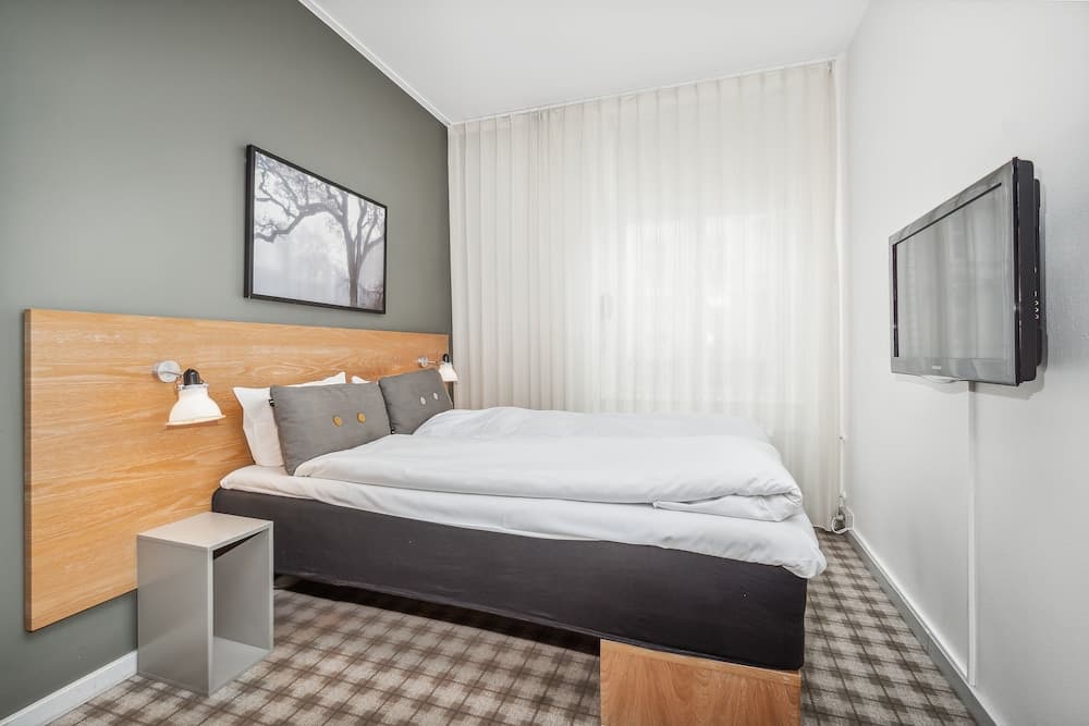 A cozy hotel room features a neatly made bed with a wooden headboard, soft lighting, a framed tree artwork, sheer curtains, and a wall-mounted TV, ideal for travelers seeking comfort and simplicity.