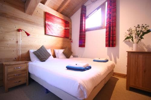A cozy attic bedroom with wooden walls, a neatly made bed adorned with blue towels, vibrant red curtains, and warm lighting creates an inviting atmosphere ideal for travelers seeking a comfortable retreat.