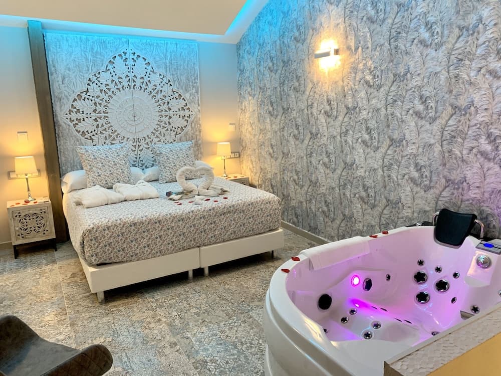 A luxurious hotel suite features a cozy bed adorned with swan towels and rose petals, a glowing hot tub with purple lights, and elegant decor, perfect for a romantic getaway.