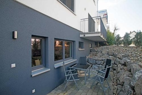 A cozy outdoor seating area beside a modern gray building with stone walls, perfect for travelers seeking a quiet retreat or a relaxing dining spot.