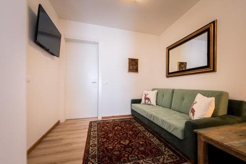 A cozy living room with a green sofa adorned with patterned cushions, a vibrant rug, and a wall-mounted TV creates an inviting atmosphere ideal for travelers seeking comfort in a modern apartment setting.