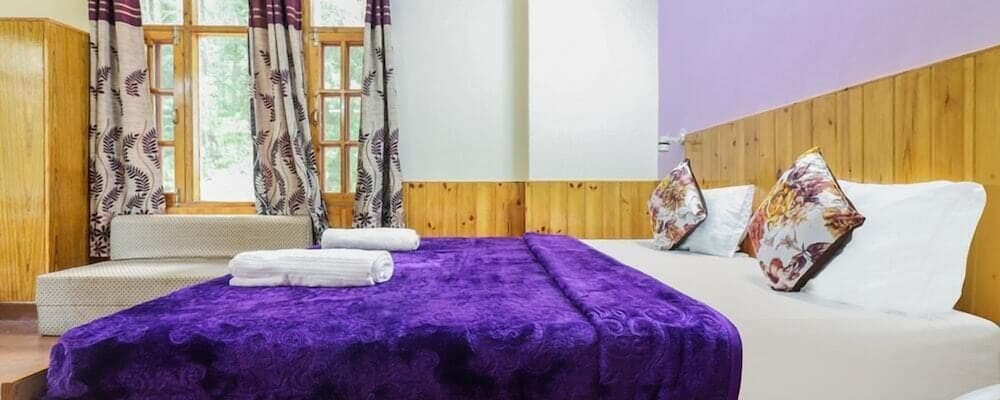 A cozy hotel room with wooden walls, purple accents, and floral curtains offers a serene retreat for travelers seeking comfort and relaxation.