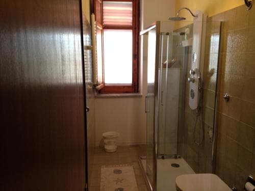 A cozy bathroom with a glass-enclosed shower, tiled walls, a small window with red shutters, and a starfish-patterned rug creates a serene and functional space ideal for travelers seeking comfort in a compact setting.