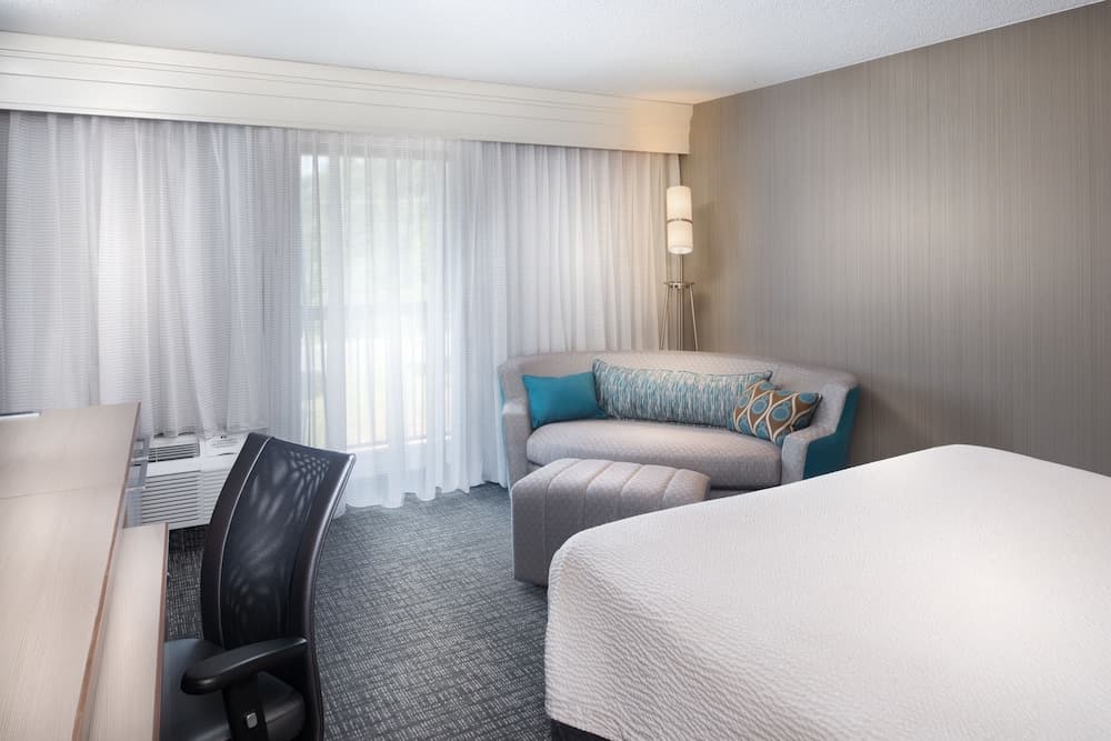 A cozy hotel room features a neatly made bed, a comfortable sofa adorned with blue and patterned pillows, a desk with an ergonomic chair, and soft natural light filtering through sheer curtains, ideal for travelers seeking relaxation and productivity.