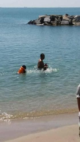 A serene beach scene captures children playing in clear turquoise waters near a rocky outcrop, evoking a peaceful seaside vacation vibe ideal for family getaways.
