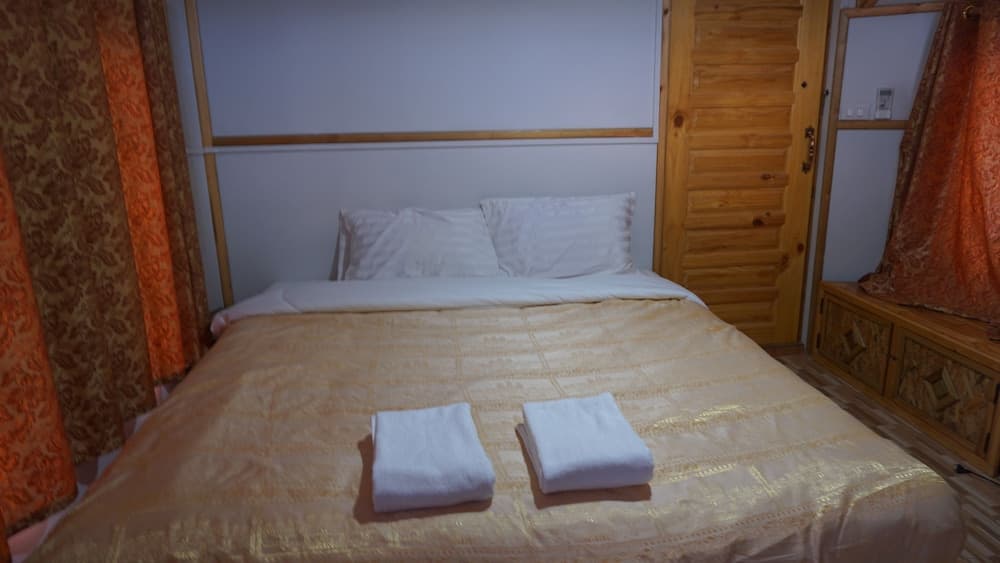 A cozy bedroom with a neatly made bed adorned in gold-patterned bedding, white towels, wooden furnishings, and warm orange curtains, ideal for travelers seeking comfort and relaxation.