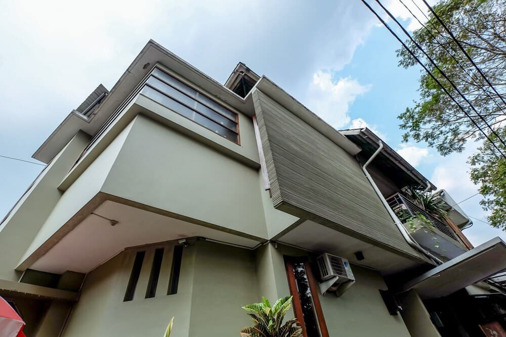 A modern two-story house with a unique architectural design, featuring large windows, a balcony adorned with greenery, and an air conditioning unit, set against a partly cloudy sky, ideal for travelers seeking a stylish urban accommodation.