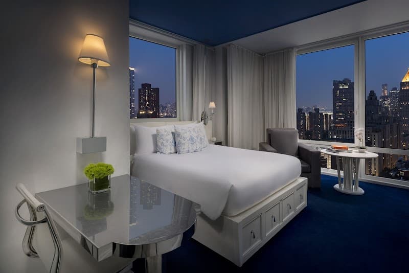 A cozy hotel room with a sleek design features large windows showcasing the vibrant New York City skyline at night, ideal for travelers seeking a luxurious urban retreat.