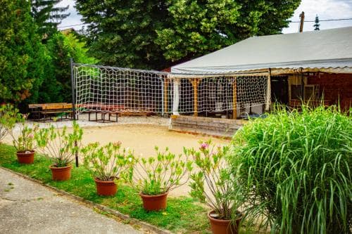A serene outdoor recreational area features a volleyball net, lush greenery, potted plants, and a shaded pavilion, ideal for family gatherings or leisurely activities in nature.