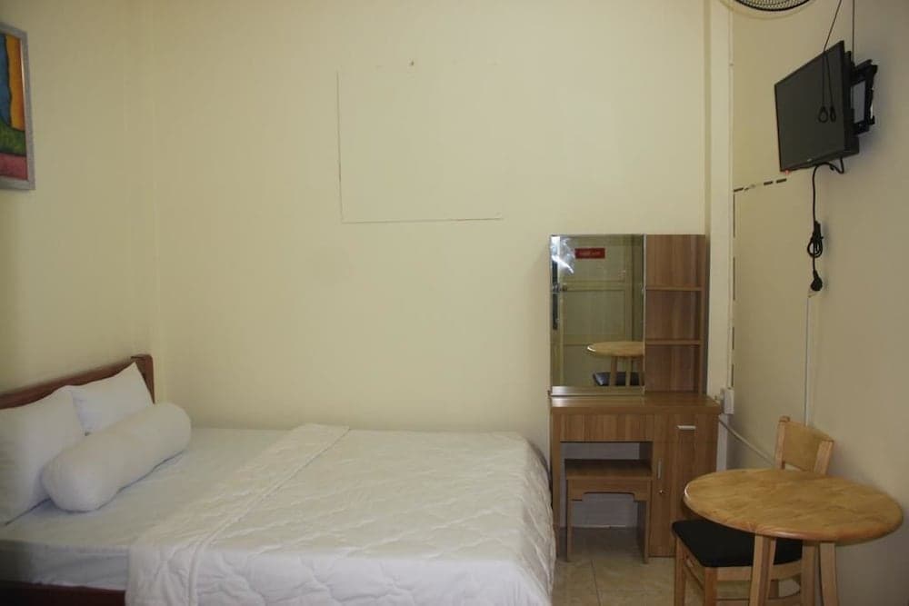 A cozy budget hotel room features a neatly made bed, wooden furniture including a desk with a mirror and a small table with chairs, a wall-mounted TV, and a minimalist decor, ideal for travelers seeking simple accommodations.