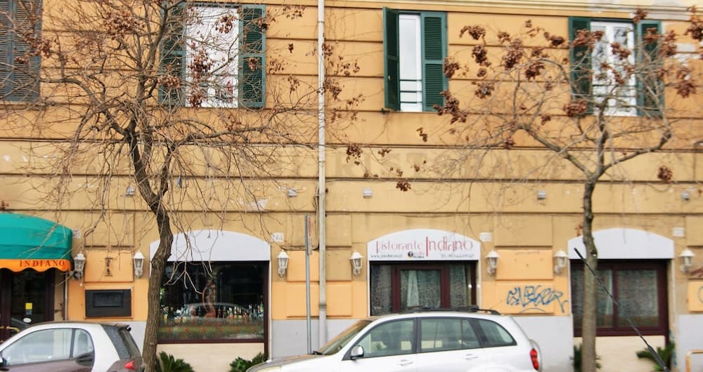 A cozy European street scene features a mustard-yellow building with green shutters, bare trees, parked cars, and a restaurant named "Ristorante Indiano," ideal for travelers seeking local dining experiences.