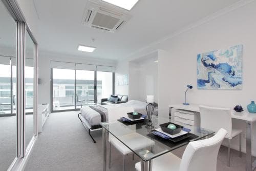 A modern, minimalist apartment features a sleek glass dining table, cozy seating area, and large windows with balcony access, creating an inviting and stylish space ideal for urban travelers seeking comfort and sophistication.