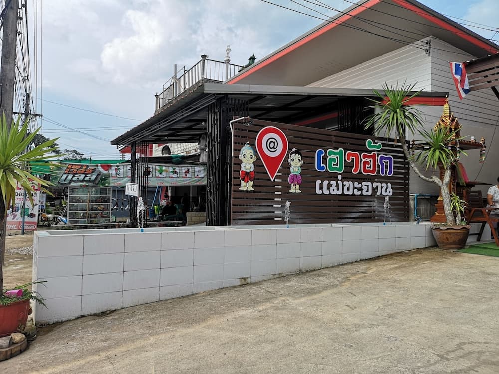 A vibrant Thai roadside eatery with colorful signage, playful cartoon characters, and tropical plants invites travelers to explore local cuisine in a casual, sunny setting.