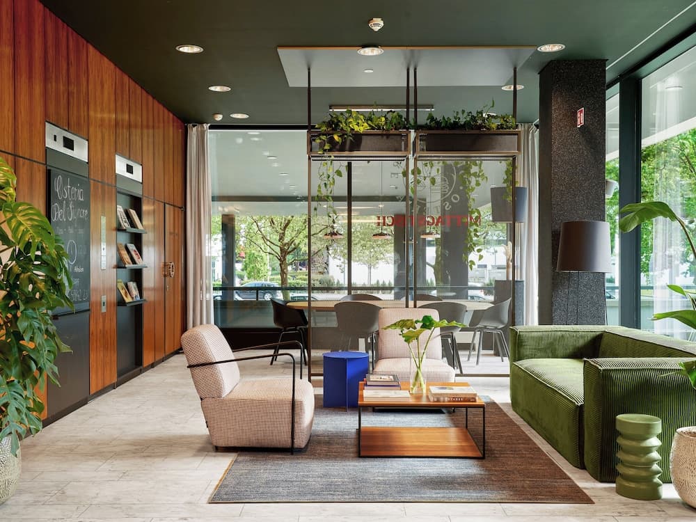 A modern hotel lobby exudes a cozy yet sophisticated vibe with its wooden paneling, lush greenery, comfortable seating, and large windows offering a serene outdoor view, ideal for travelers seeking relaxation and style.