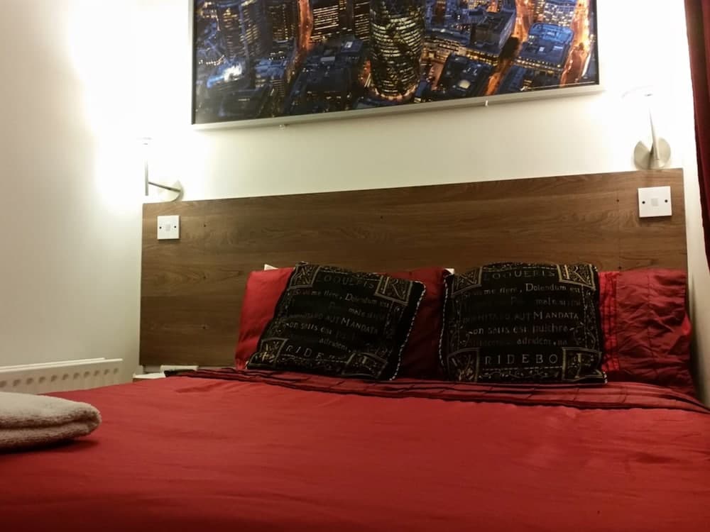 A cozy bedroom features a red bed adorned with patterned pillows, a wooden headboard, wall-mounted lights, and a vibrant cityscape artwork, ideal for travelers seeking a warm and stylish accommodation.