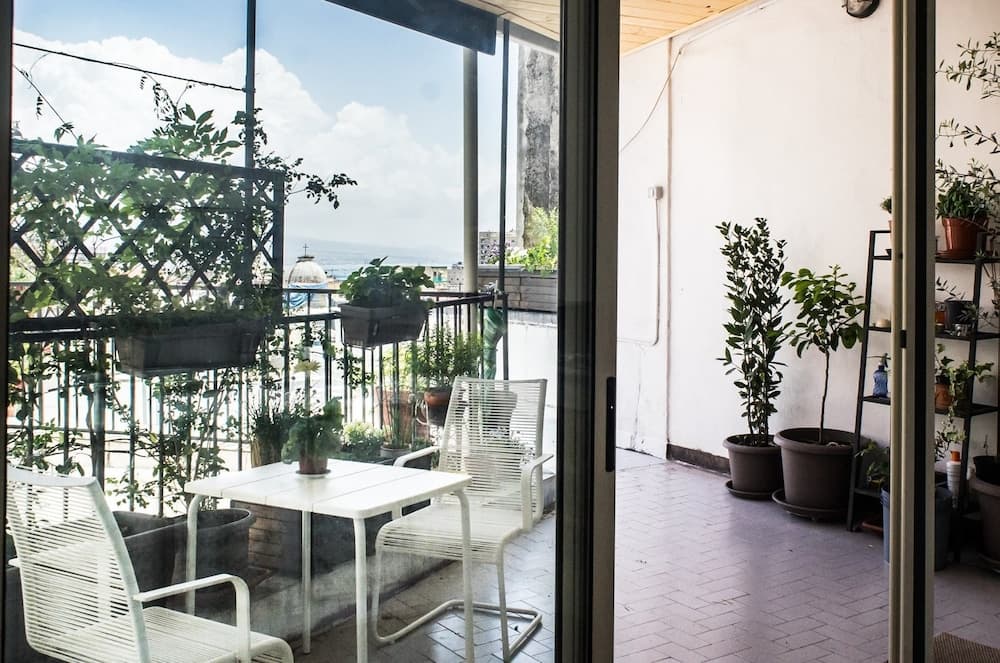 A serene balcony with white furniture and lush potted plants overlooks a scenic coastal view, ideal for travelers seeking a peaceful retreat.