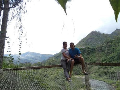 Two travelers relax on a wooden bridge with netting, enjoying the serene mountainous landscape, ideal for adventure seekers exploring lush, green terrains.