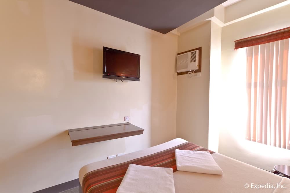 A cozy hotel room features a neatly made bed with folded towels, a wall-mounted TV, a floating desk, and a bright window with blinds, ideal for travelers seeking comfort and simplicity.
