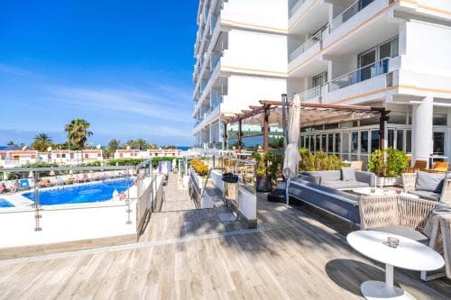 A sunny seaside terrace features comfortable seating, lush greenery, and a vibrant poolside view, ideal for travelers seeking relaxation at a beachfront resort.