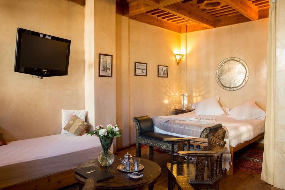 A cozy hotel room with warm beige walls, wooden beams, and traditional decor creates an inviting atmosphere, featuring a comfortable bed, ornate furniture, and a flat-screen TV, perfect for travelers seeking relaxation and cultural ambiance.