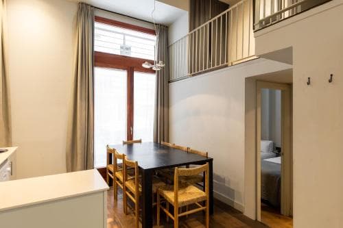A cozy apartment interior features a modern dining area with wooden chairs, a sleek black table, and a lofted bedroom above, ideal for travelers seeking a stylish yet comfortable urban stay.