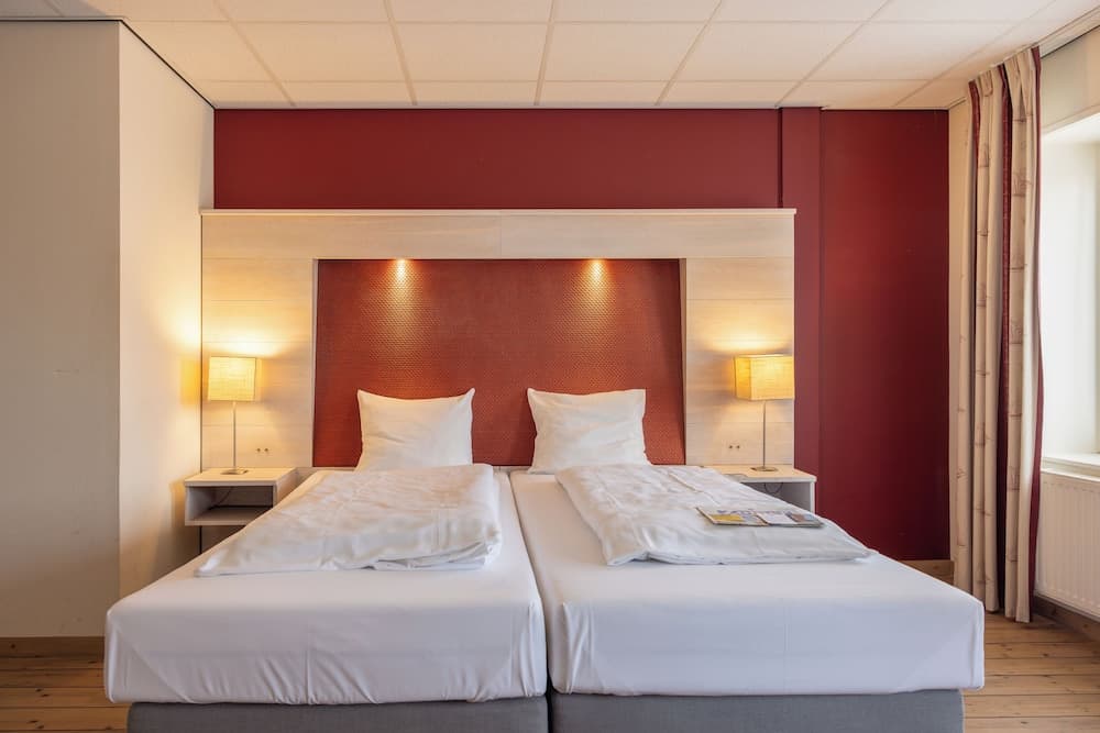 A cozy hotel room features twin beds with white linens against a warm red accent wall, flanked by bedside lamps, creating an inviting atmosphere ideal for travelers seeking comfort and relaxation.