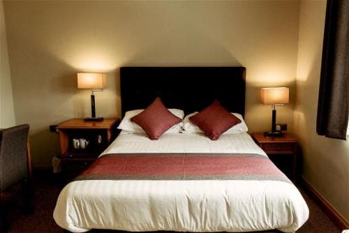 A cozy hotel room features a neatly made bed with red accents, warm bedside lamps, and a calm ambiance, ideal for travelers seeking comfort and relaxation.