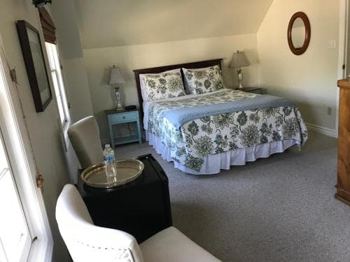 A cozy, well-lit guest room features a neatly made bed with floral bedding, white chairs, a small table with refreshments, and soft lighting, ideal for travelers seeking comfort and relaxation in a charming accommodation setting.