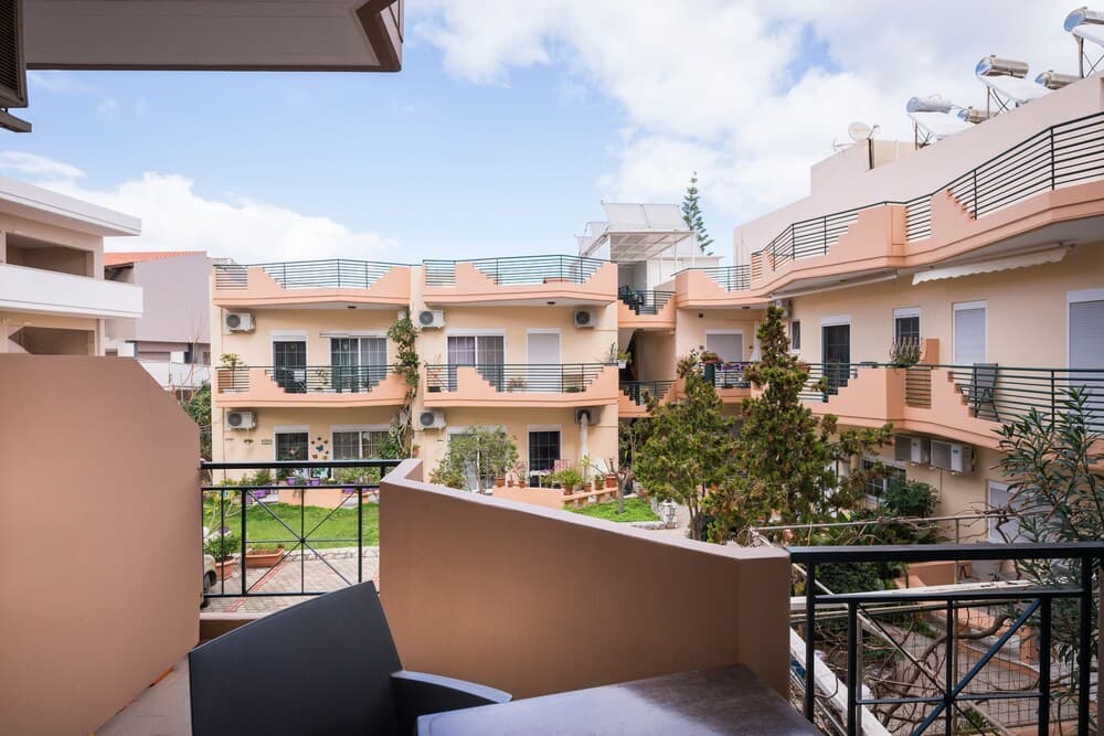 A serene residential courtyard with pastel-colored buildings, balconies adorned with plants, and a clear blue sky creates an inviting atmosphere ideal for leisurely stays or vacation rentals.