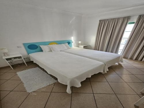 A cozy hotel room features two neatly made beds with a colorful headboard, beige curtains framing a balcony door, and tiled flooring, ideal for travelers seeking comfort and relaxation.
