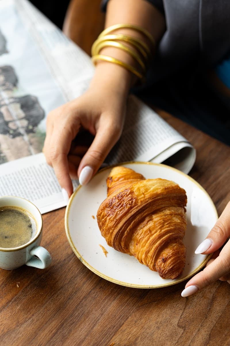 A cozy café scene in Paris features hands adorned with gold bangles reaching for a flaky croissant on a white plate, accompanied by a steaming cup of coffee and an open newspaper, perfect for a leisurely morning travel experience.
