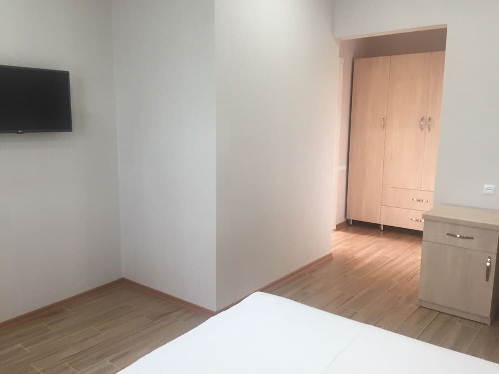 A minimalist bedroom with white walls, wooden flooring, a mounted TV, and light wood furniture exudes a serene and cozy atmosphere, ideal for travelers seeking a clean and simple accommodation option.