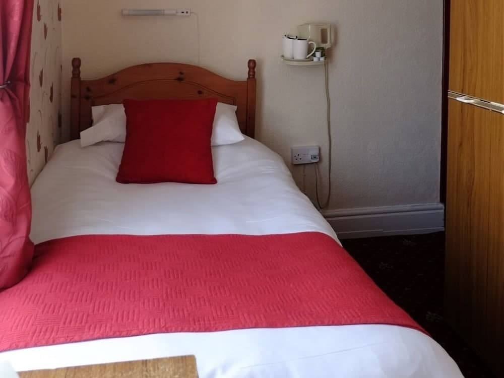 A cozy hotel room features a neatly made bed with white linens and a red throw pillow, complemented by a wooden headboard, patterned curtains, and a wall-mounted shelf with tea cups, ideal for travelers seeking comfort and simplicity.