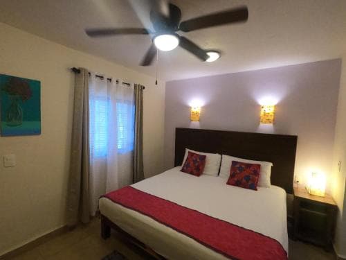 A cozy hotel room features a neatly made bed with red accents, soft lighting from wall sconces, and a ceiling fan, creating a warm and inviting atmosphere ideal for travelers seeking relaxation.