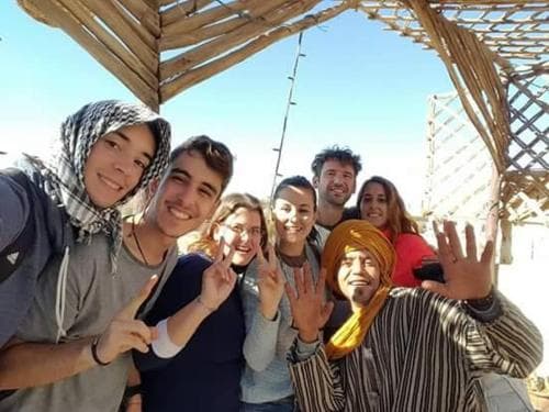 A cheerful group of travelers poses under a rustic wooden shelter in a sunny desert setting, showcasing cultural attire and friendly gestures ideal for adventure tourism content.