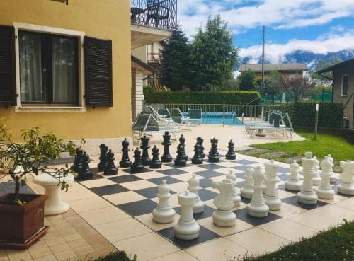 A serene outdoor patio features a giant chess set on a tiled floor, surrounded by lush greenery, a swimming pool, and distant mountains, ideal for leisurely stays at a cozy vacation rental.