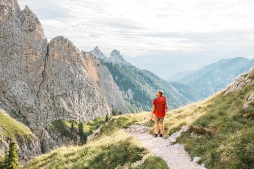 A hiker in a red jacket stands on a rugged mountain trail overlooking dramatic peaks and lush valleys, capturing the adventurous spirit of outdoor exploration ideal for trekking enthusiasts.