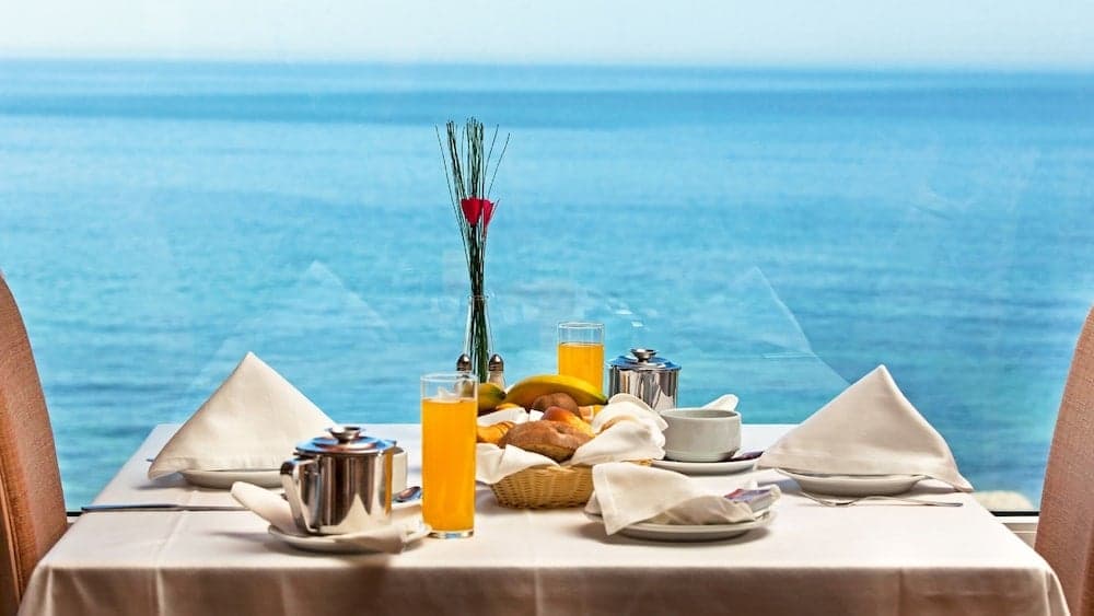 A serene coastal dining setup features a beautifully arranged breakfast table with orange juice, bread, and tea, set against the tranquil backdrop of the ocean, ideal for travelers seeking a relaxing seaside retreat experience.