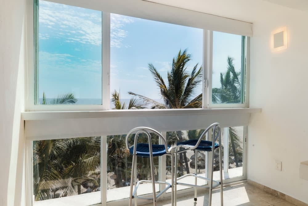 A serene coastal retreat features a minimalist indoor space with blue bar stools by a large window, offering a breathtaking view of palm trees and the ocean, ideal for vacation rental listings.