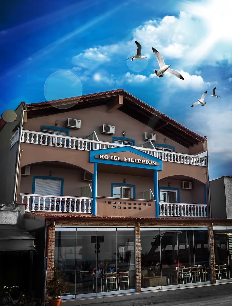 A charming hotel with blue accents and balconies stands under a vibrant sky dotted with seagulls, inviting travelers to enjoy its serene seaside ambiance.