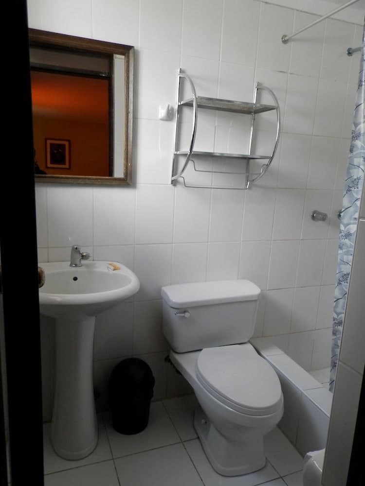 A compact bathroom with white tiled walls features a pedestal sink, toilet, metal shelving, and a floral shower curtain, ideal for travelers seeking efficient space utilization in accommodations.
