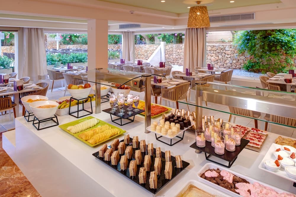 A vibrant indoor buffet area with an array of colorful desserts, fresh fruits, and pastries set against a backdrop of elegant dining tables and lush greenery outside, ideal for travelers seeking a luxurious dining experience in a resort setting.