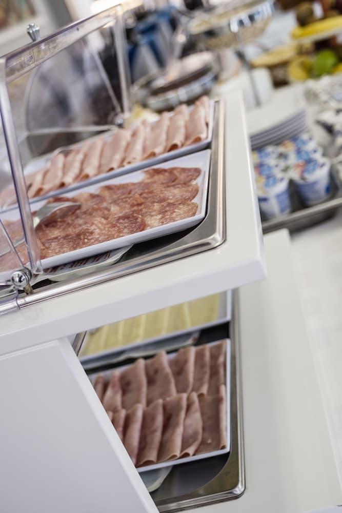 A well-lit hotel buffet station showcases neatly arranged slices of ham and prosciutto under glass covers, inviting travelers to enjoy a fresh breakfast spread.