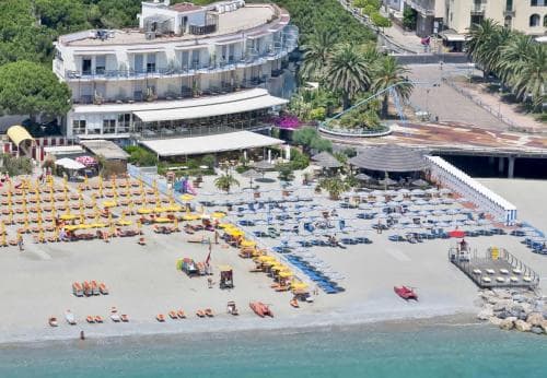 A vibrant beach scene with colorful umbrellas and lounge chairs lines the sandy shore beside turquoise waters, set against a backdrop of palm trees and a bustling coastal town, ideal for a relaxing seaside getaway.